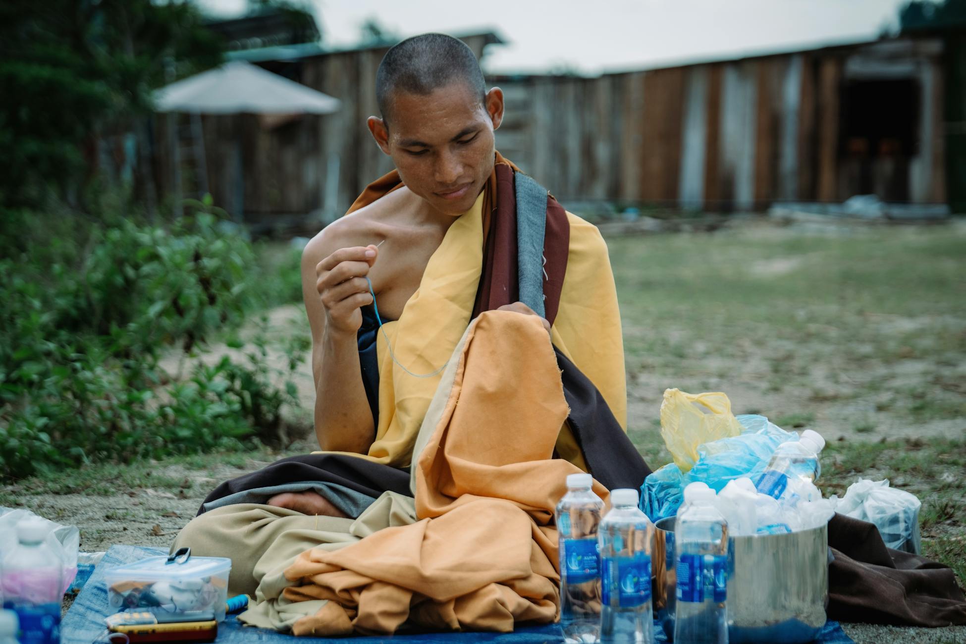 A thoughtful monk in traditional attire sits meditatively surrounded by bottles in an outdoor setting.