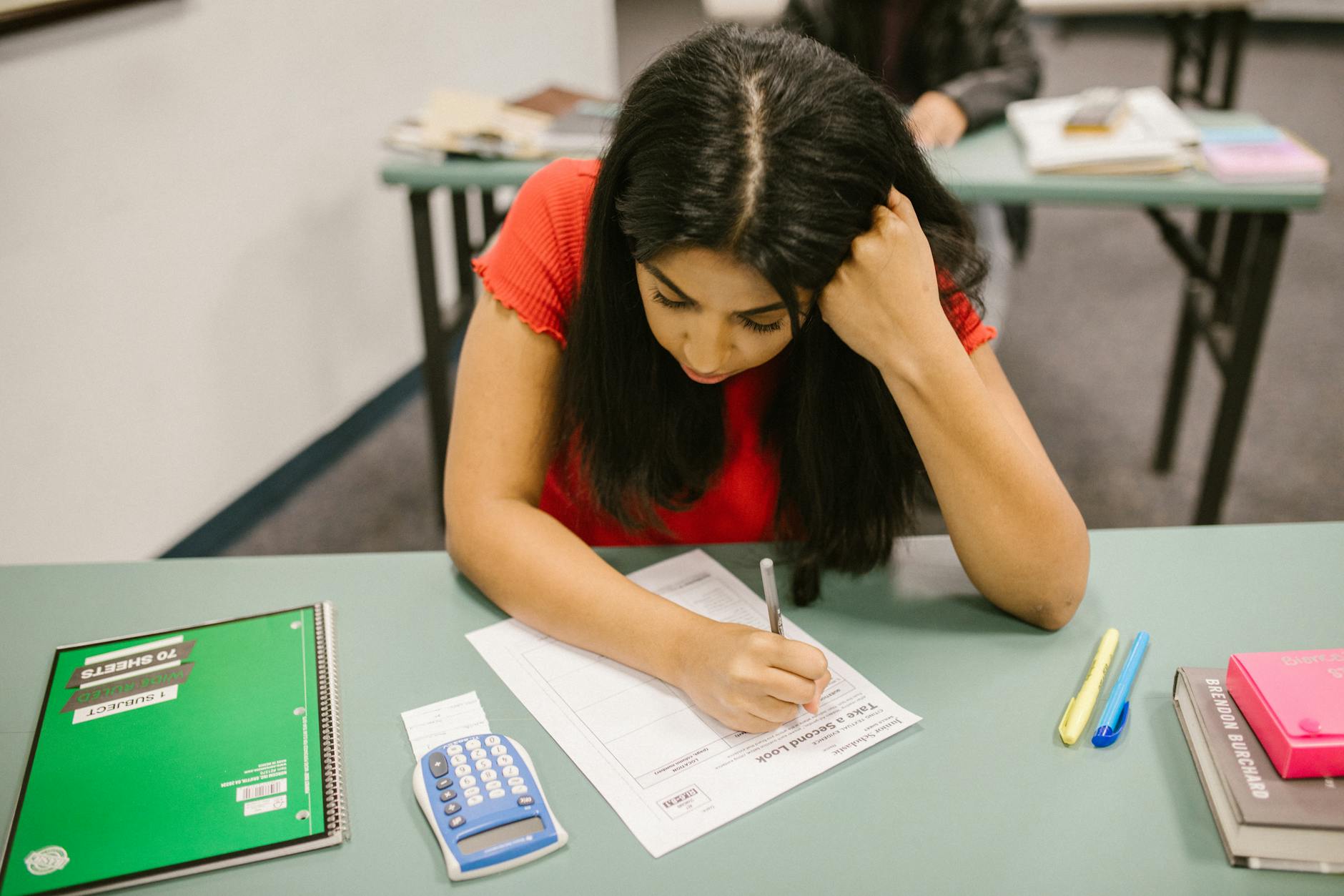 Young man looking frustrated while sitting at a desk with exam papers in a classroom.