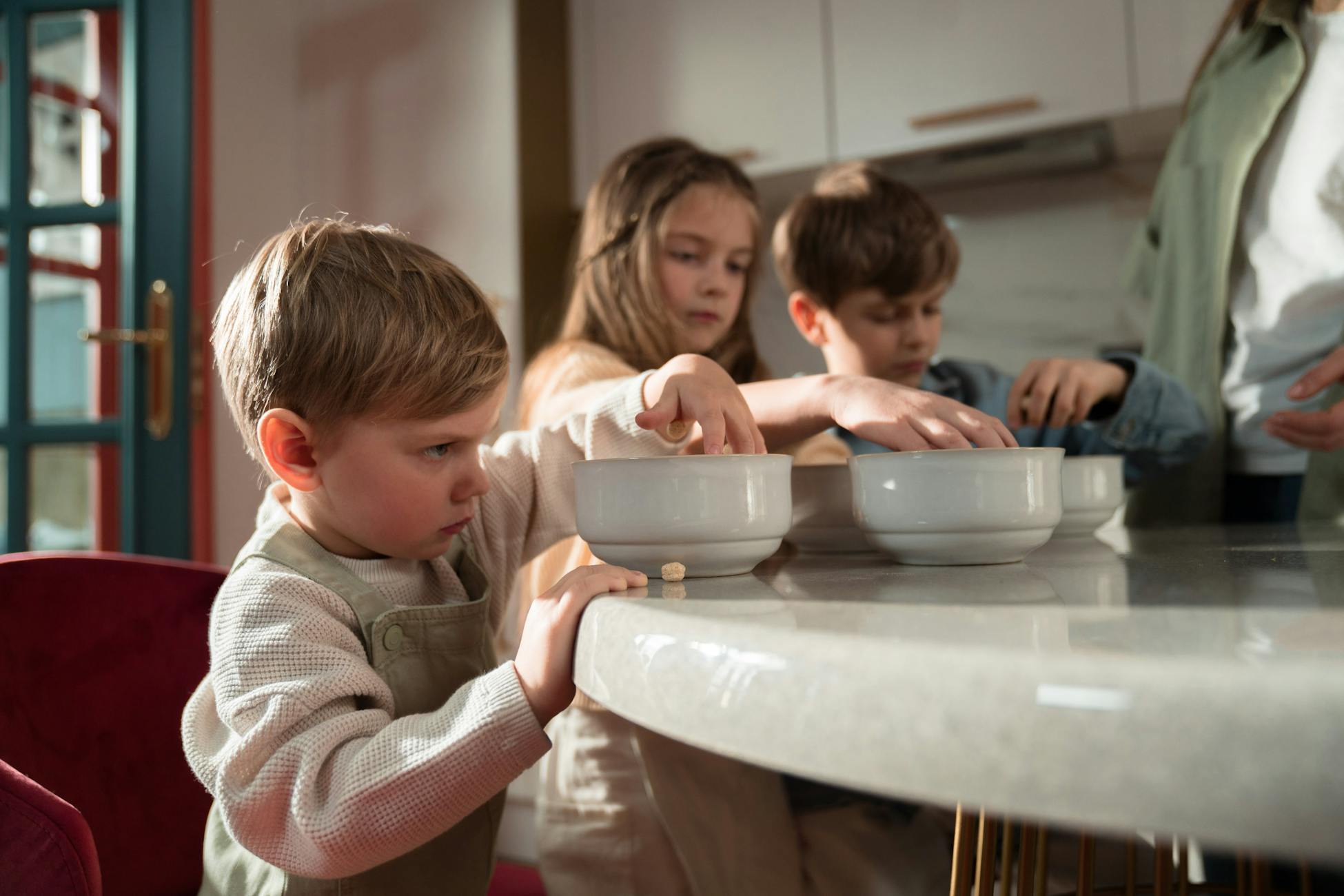 Three children gather around a table enjoying breakfast indoors on a sunny morning.